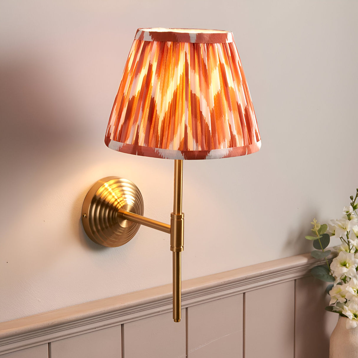 Endon Castleigh wall light with brushed aged brass T-shaped arm and hand-pleated apricot orange Zigzag faux silk shade, mounted on a panelled hallway wall.