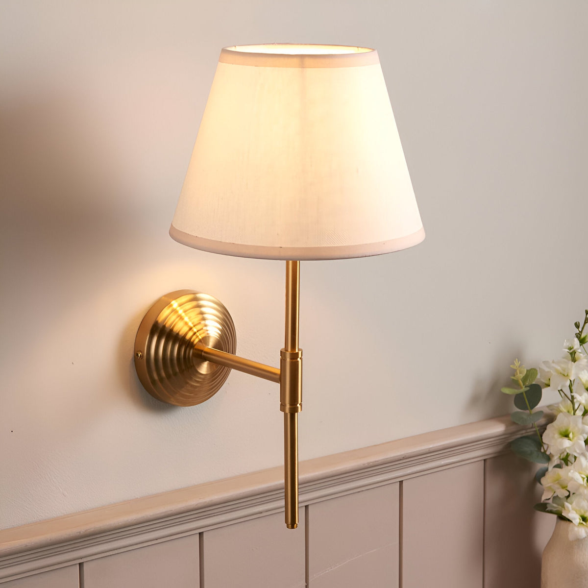 Castleigh Ivy Wall Light in antique brass with a tapered vintage white fabric shade, mounted on a beige wall in a hallway setting.