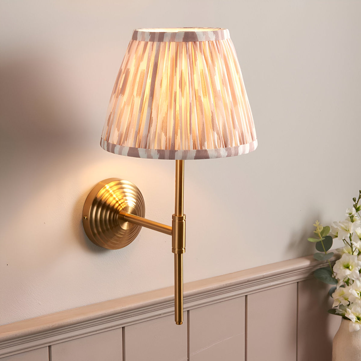 Endon Castleigh wall light with brushed aged brass stepped backplate, adjustable T-arm and cream ikat pleated faux silk tapered shade, lit against a pale wall.