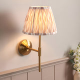 Endon Castleigh wall light with brushed aged brass stepped backplate, adjustable T-arm and cream ikat pleated faux silk tapered shade, lit against a pale wall.