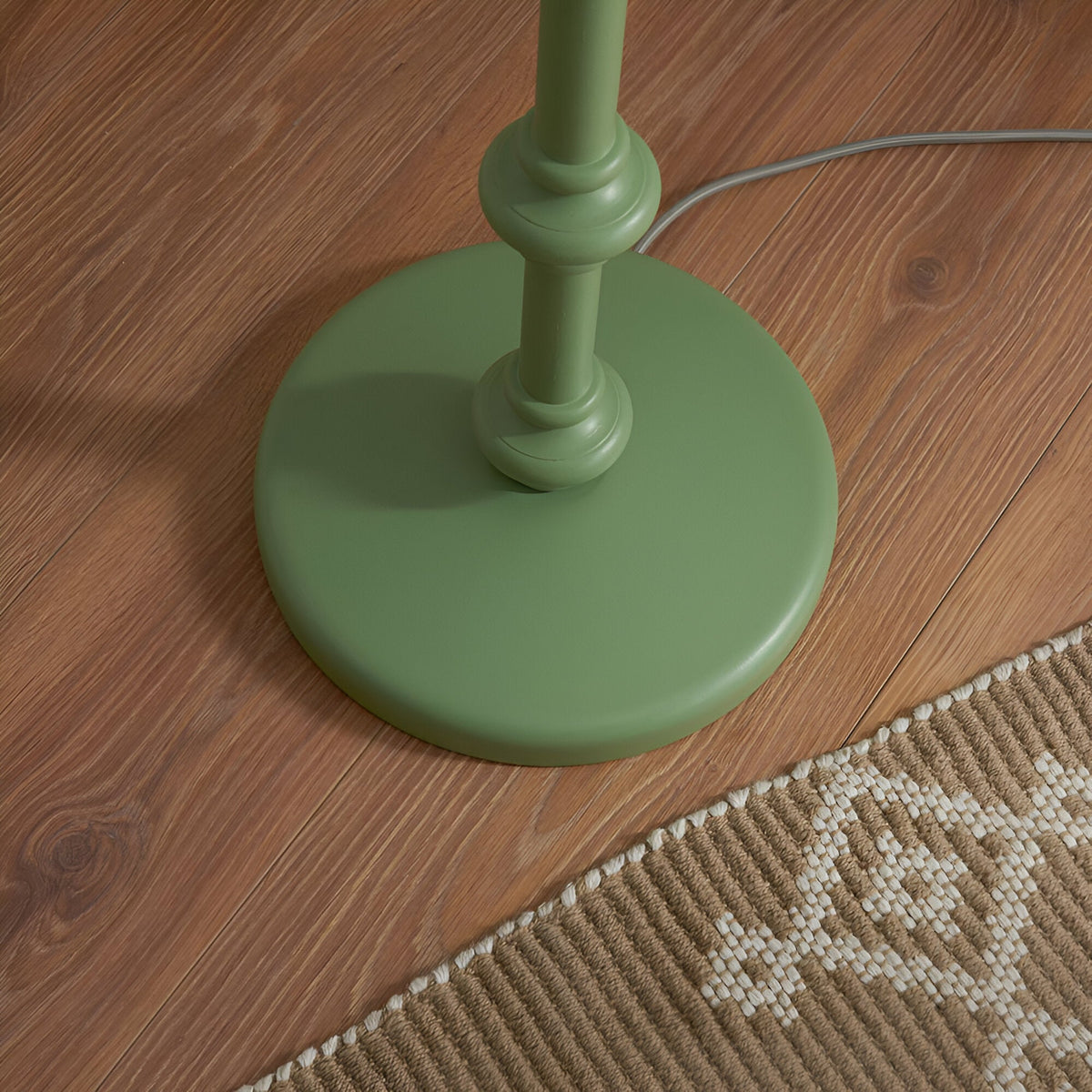 Close-up of the Endon Bevbury floor lamp's green turned wood spindle base on wooden flooring beside a woven rug, with grey braided flex trailing.