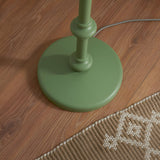 Close-up of the Endon Bevbury floor lamp's green turned wood spindle base on wooden flooring beside a woven rug, with grey braided flex trailing.