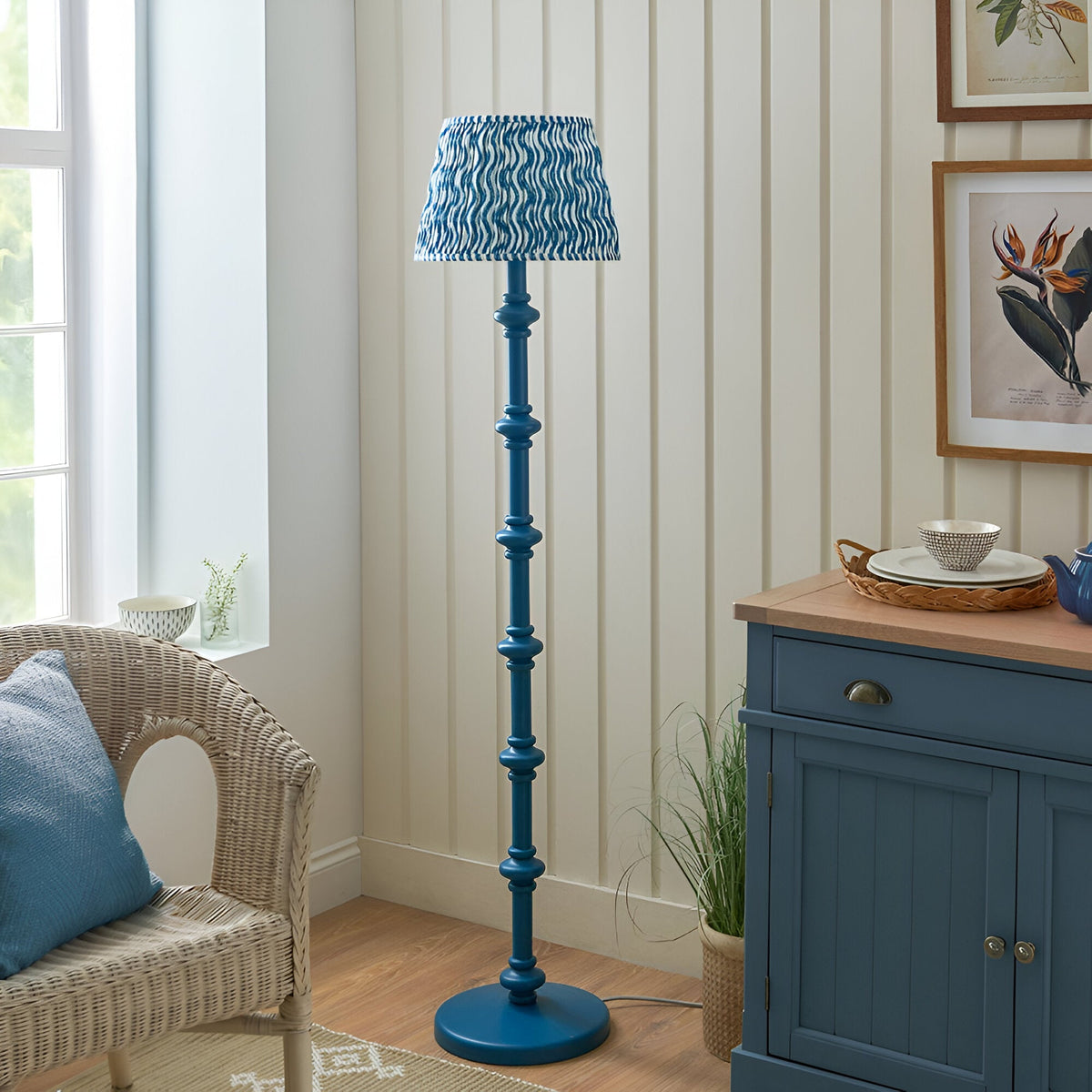 Endon Bevbury Ripple floor lamp with blue turned wood spindle base and blue wave-patterned pleated shade, beside a wicker chair and blue sideboard.