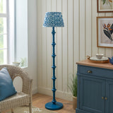 Endon Bevbury Ripple floor lamp with blue turned wood spindle base and blue wave-patterned pleated shade, beside a wicker chair and blue sideboard.