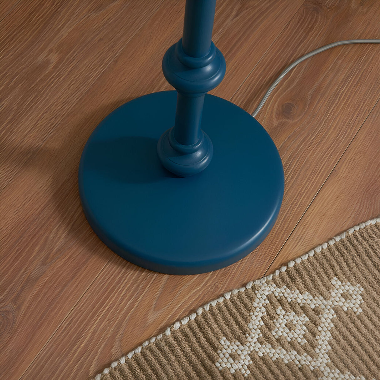 Close-up of the Endon Bevbury Ripple floor lamp's marlin blue turned wooden spindle base with grey braided flex on wood flooring beside a woven rug.