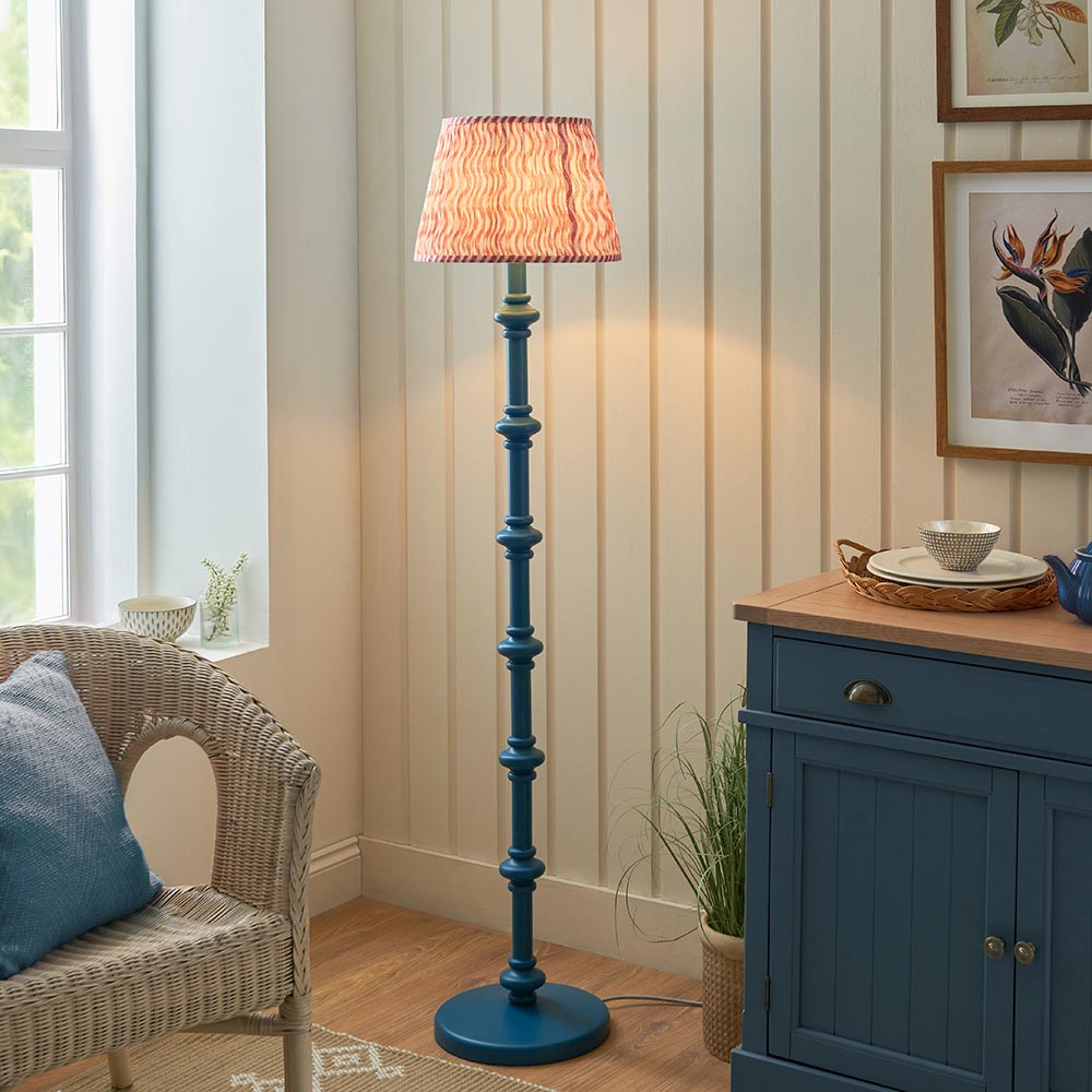 Endon Bevbury Ripple floor lamp with a blue turned wood spindle base and pink patterned pleated shade, beside a wicker armchair and blue sideboard.