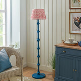Endon Bevbury Ripple floor lamp with blue turned wood spindle base and pink patterned pleated shade, beside a rattan chair and blue sideboard.