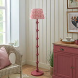 Endon Bevbury Ripple floor lamp with pink turned wood spindle base and ripple pleated shade, styled beside a wicker chair and pink sideboard.