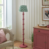 Endon Bevbury floor lamp with pink clay turned wood spindle base and green zigzag pleated shade, styled beside a wicker chair and pink sideboard.