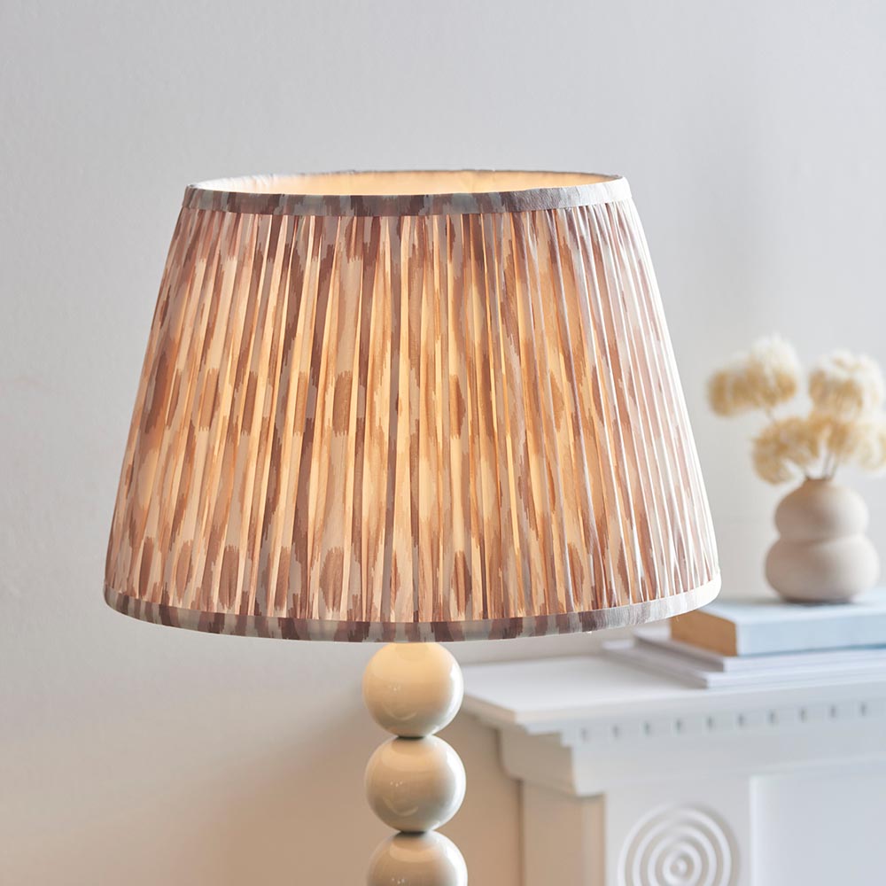 Endon Brenleigh floor lamp's white and natural Ikat pleated shade shown lit on a glossy cream stacked sphere base beside a mantel with dried flowers.