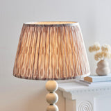 Endon Brenleigh floor lamp's white and natural Ikat pleated shade shown lit on a glossy cream stacked sphere base beside a mantel with dried flowers.