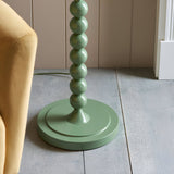 Close-up of the Endon Brenleigh Ripple floor lamp base, showing stacked green gloss spheres on a round base beside a mustard chair on pale wood flooring.