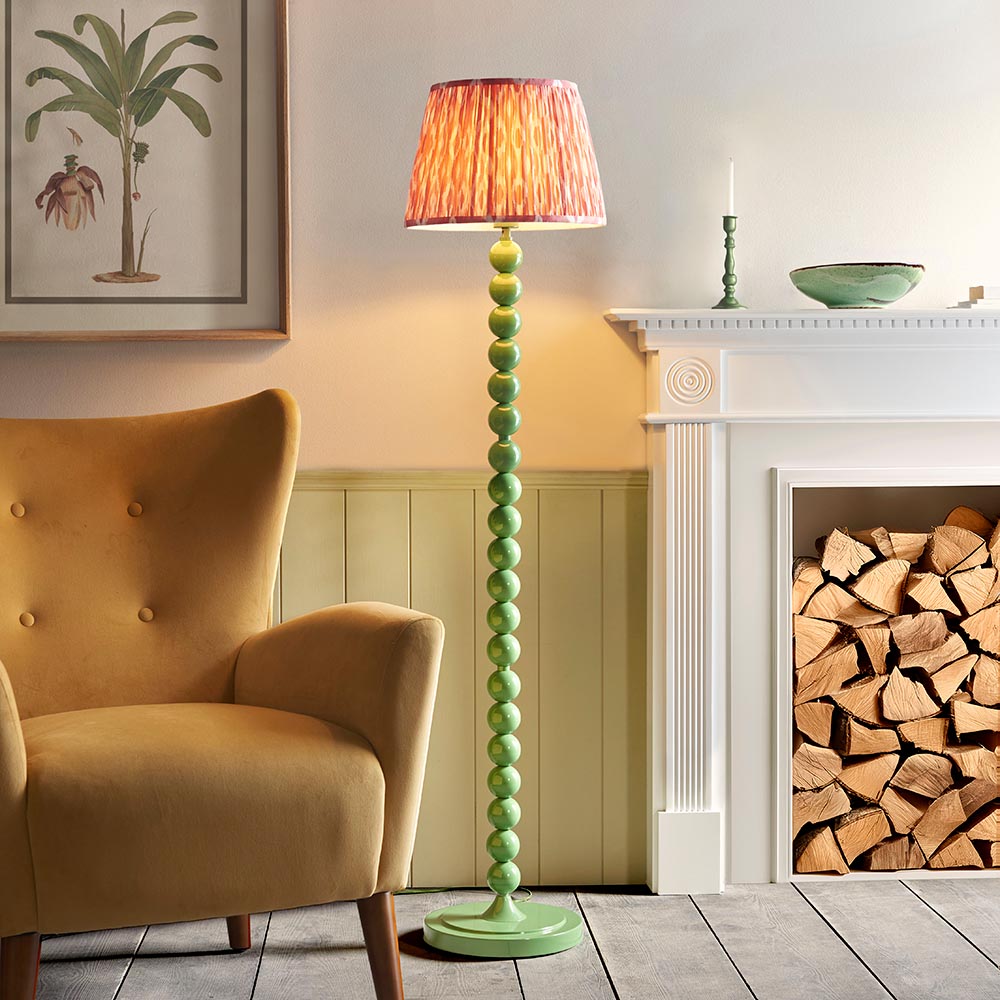 Brenleigh Ikat Floor Lamp with stacked glossy green sphere base and pink patterned pleated fabric shade, shown beside a fireplace in a living room.