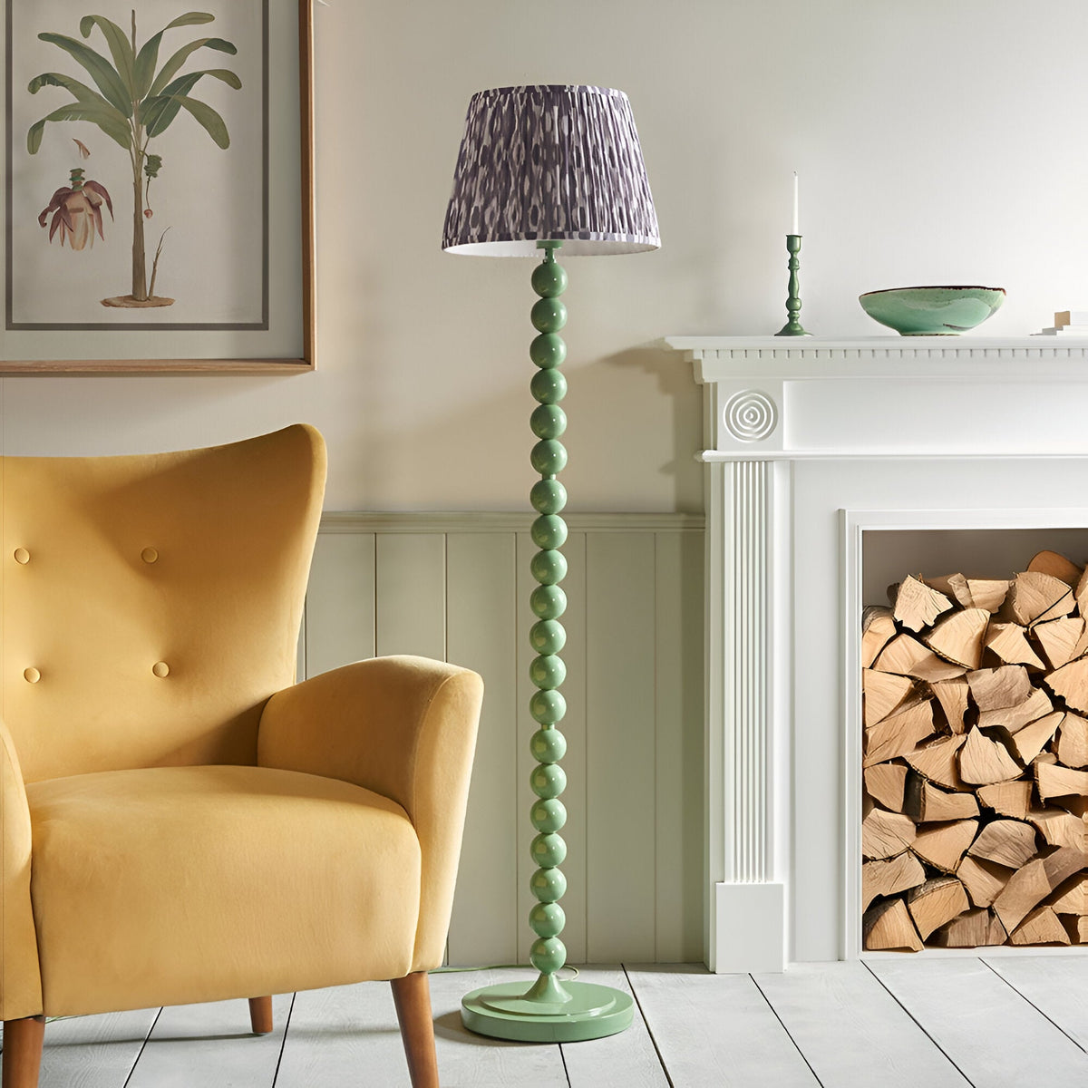 Endon Brenleigh floor lamp with stacked green gloss sphere base and grey Ikat pleated shade, beside a yellow armchair and white fireplace with logs.
