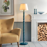 Brenleigh Ivy Floor Lamp with stacked blue gloss sphere base and white fabric tapered shade, shown lit beside a mustard armchair and fireplace.