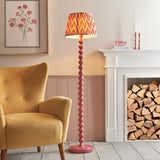 Brenleigh Zigzag Floor Lamp in pink with stacked sphere metal base and pleated orange zigzag fabric shade, shown beside a mustard armchair and fireplace.