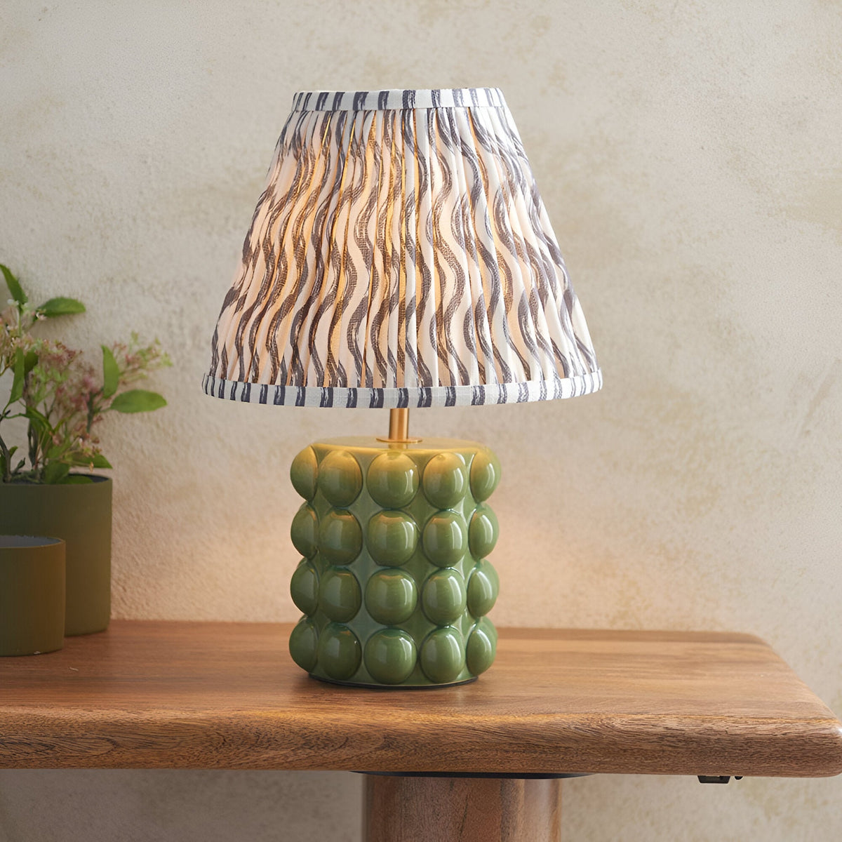 Endon Amberley Ripple table lamp with green ceramic bobble base, brass stem and pleated grey wave-print shade on a wooden console beside a plant.