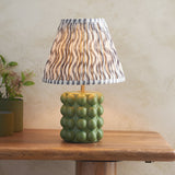 Endon Amberley Ripple table lamp with green ceramic bobble base, brass stem and pleated grey wave-print shade on a wooden console beside a plant.