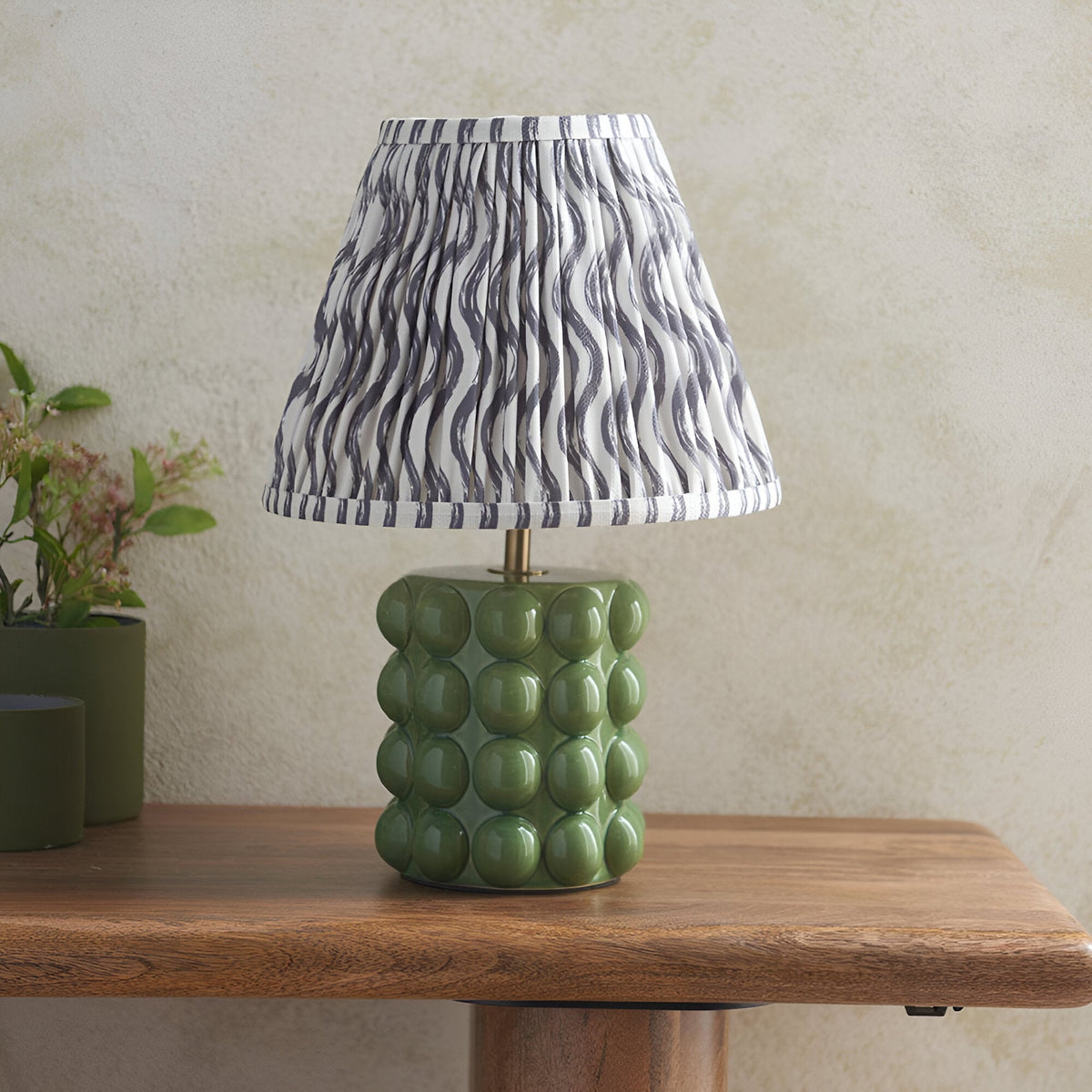 Endon Amberley Ripple table lamp with olive green ceramic bobble base and pleated grey wave-pattern shade, on a wooden table beside a potted plant.