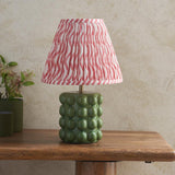 Amberley Ripple Table Lamp with a green ceramic bobble base and pink and white wavy-striped pleated shade, shown on a wooden table beside potted plants.
