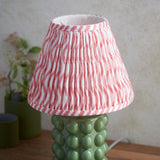 Amberley Ripple Table Lamp with green ceramic bobble base and pink-and-white ripple pattern pleated shade, shown beside potted plants on a wooden surface.