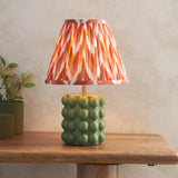 Endon Amberley table lamp with glossy olive green ceramic bobble base and pleated apricot zigzag shade, on a wooden table beside a potted plant.