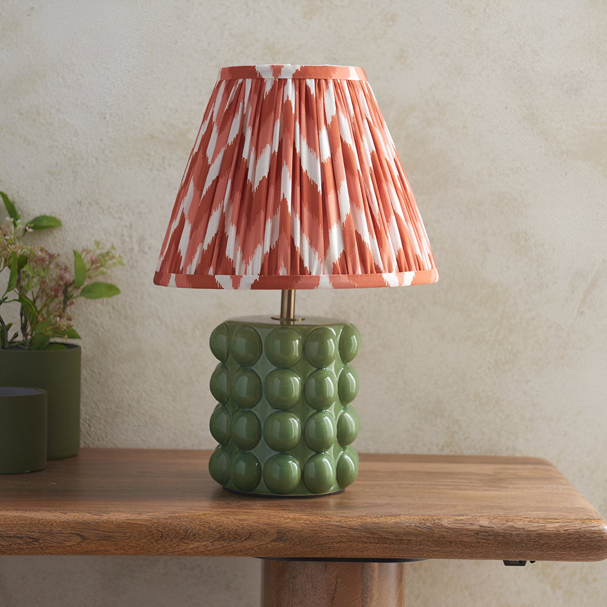 Endon Amberley table lamp with a glossy olive green bobble ceramic base and pleated apricot zigzag shade, on a wooden side table.