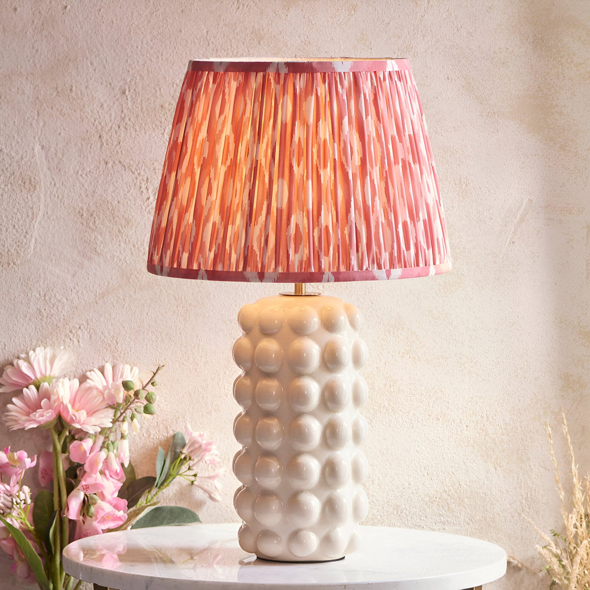 Amberley Ikat table lamp with a bobble-textured white ceramic base and pleated pink, orange and grey Ikat-print shade on a marble table.
