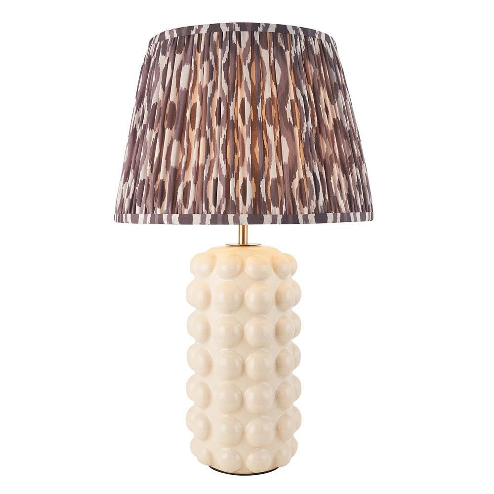 Amberley Ikat table lamp with a bobble-textured cream ceramic base, brass stem, and pleated grey-brown ikat print shade on a white background.