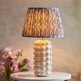 Amberley Ikat table lamp with a white ceramic bobble base and pleated grey and brown Ikat print shade, shown lit on a marble side table.