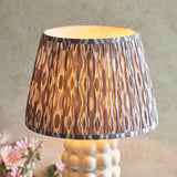 Amberley Ikat table lamp with a pleated brown, grey and white patterned Ikat shade on a white ceramic bobble base, shown lit against a neutral background.