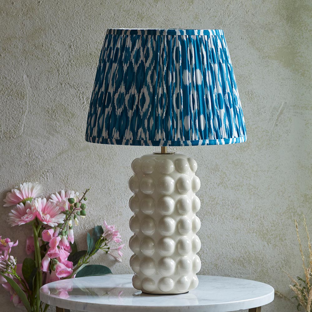 Endon Amberley Ikat table lamp with white ceramic bobble base and pleated blue and white ikat shade, on a marble table beside pink flowers.