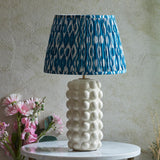 Endon Amberley Ikat table lamp with white ceramic bobble base and pleated blue and white ikat shade, on a marble table beside pink flowers.