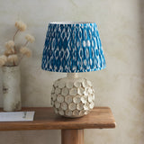 Ashbury Ikat Table Lamp with pleated blue and white patterned shade on a round ivory crackle-glaze ceramic base, styled on a wooden side table.