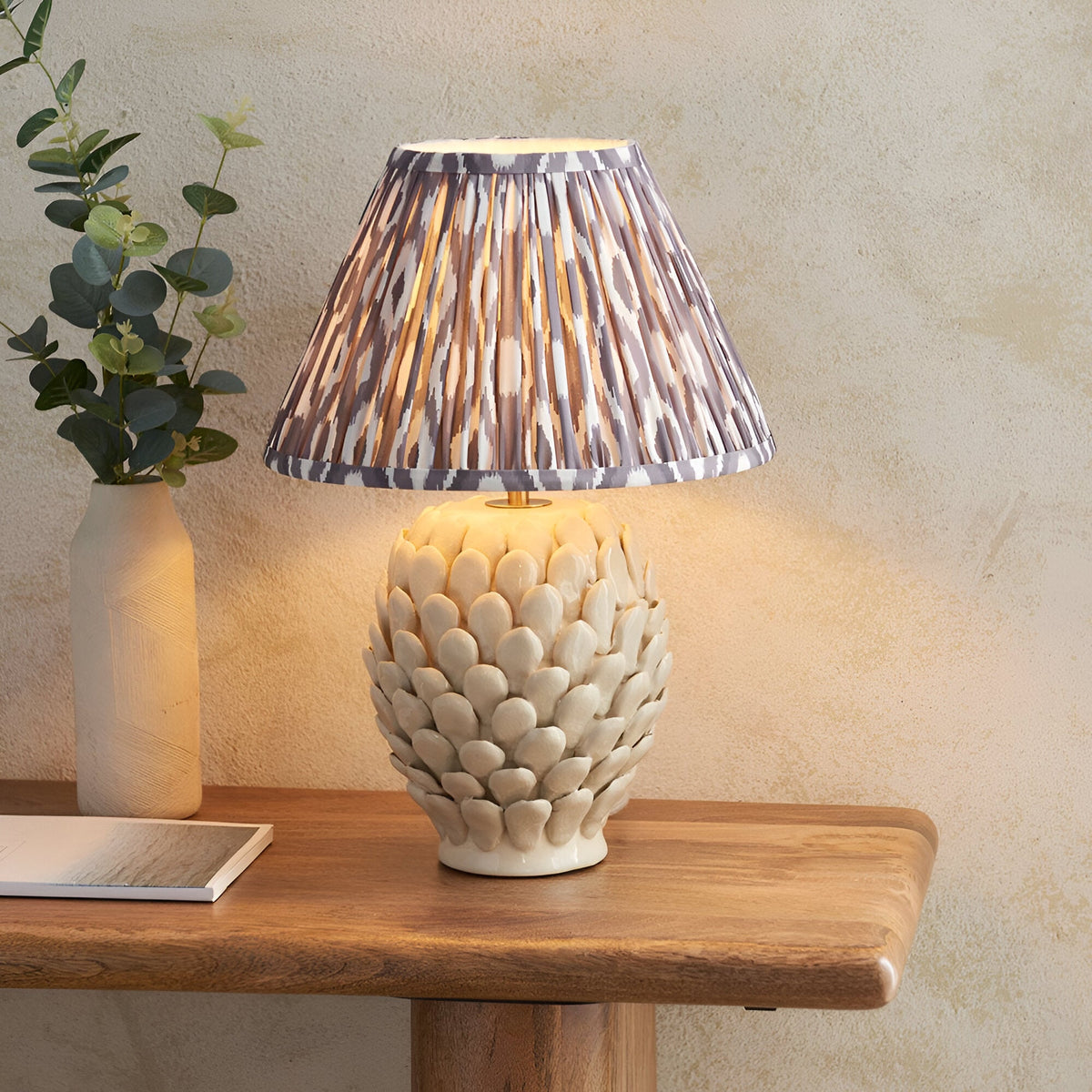 Endon Caldford Ikat table lamp lit on a wooden console, ivory ceramic leaf-detail base with pleated grey Ikat shade beside a eucalyptus vase.
