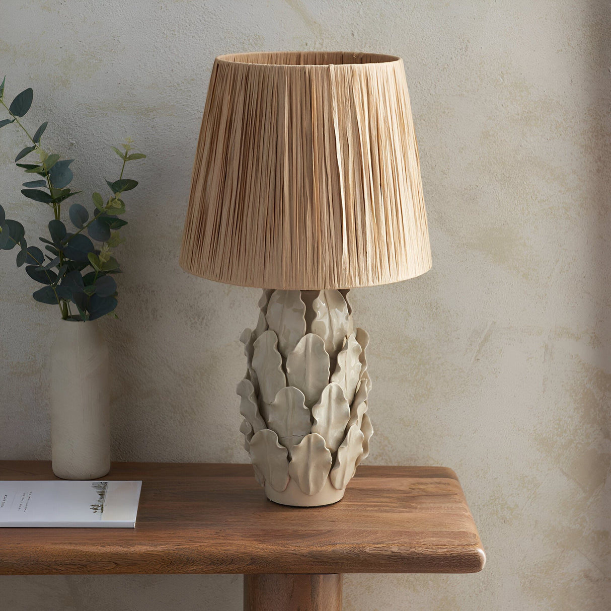 Endon Caldford table lamp with ivory crackle-glazed ceramic leaf base and natural raffia tapered shade on a wooden console beside a eucalyptus vase.