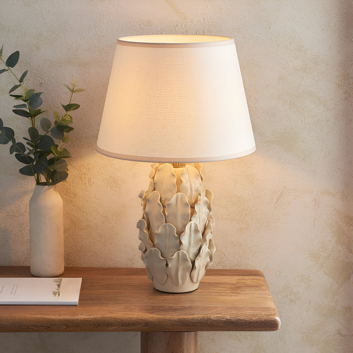 Caldford Ivy 35cm Table Lamp in ivory with ceramic leaf-textured base and white fabric shade, shown lit on a wooden console table beside a vase.