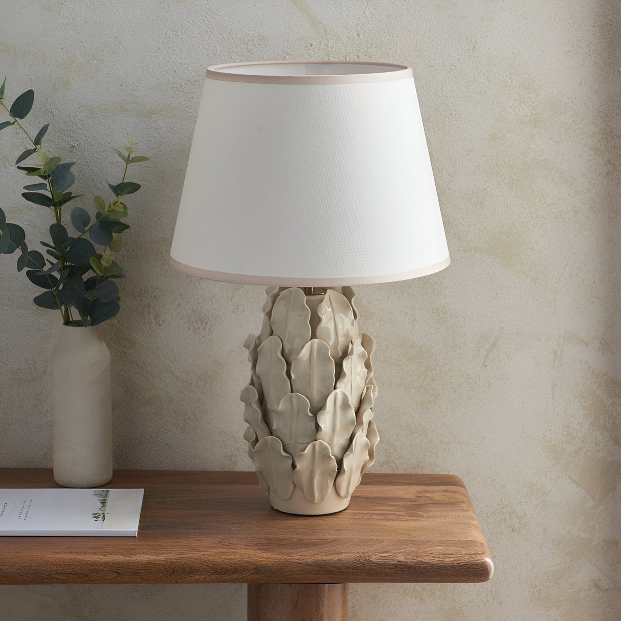 Caldford Ivy 35cm table lamp with ivory ceramic leaf-detail base and white fabric shade, styled on a wooden table beside a vase and books.
