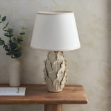 Caldford Ivy 35cm table lamp with ivory ceramic leaf-detail base and white fabric shade, styled on a wooden table beside a vase and books.