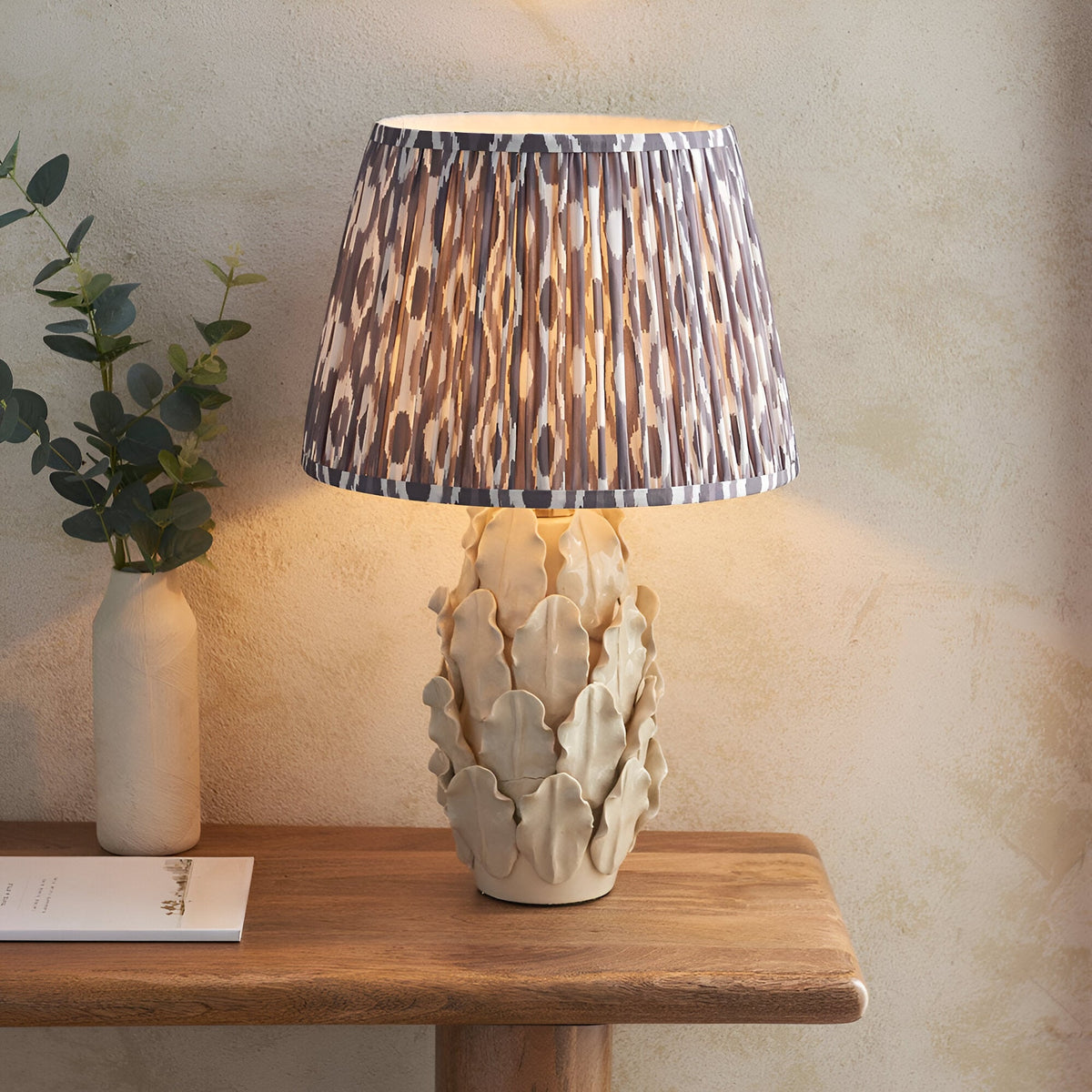 Endon Caldford Ikat 35cm table lamp with ivory crackle-glaze ceramic leaf base and pleated grey Ikat shade, on a wooden console beside a eucalyptus vase.