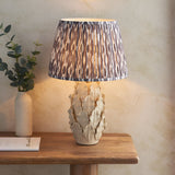 Endon Caldford Ikat 35cm table lamp with ivory crackle-glaze ceramic leaf base and pleated grey Ikat shade, on a wooden console beside a eucalyptus vase.