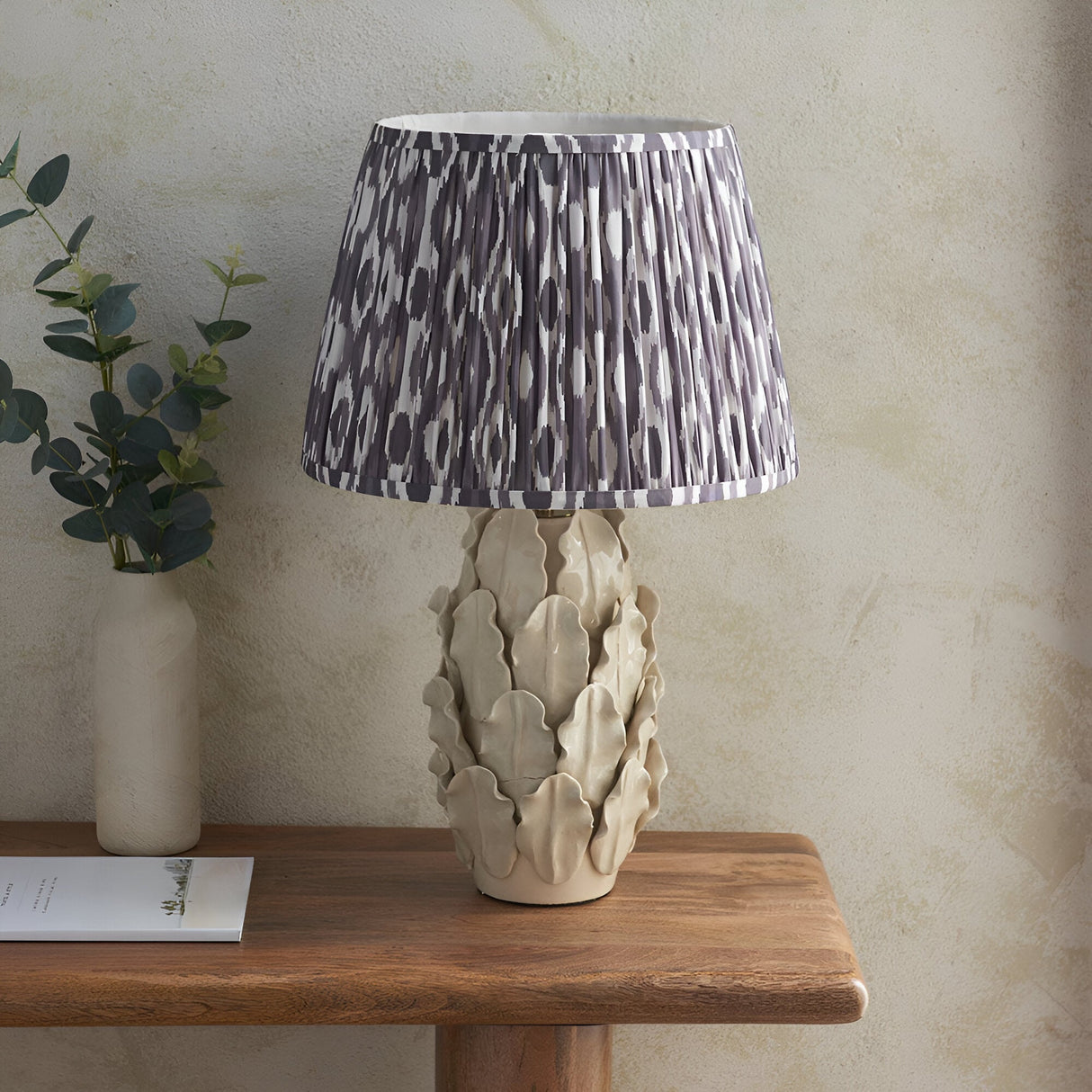 Endon Caldford Ikat 35cm table lamp with ivory crackle-glaze ceramic leaf base and grey pleated Ikat shade, on a wooden console with eucalyptus.