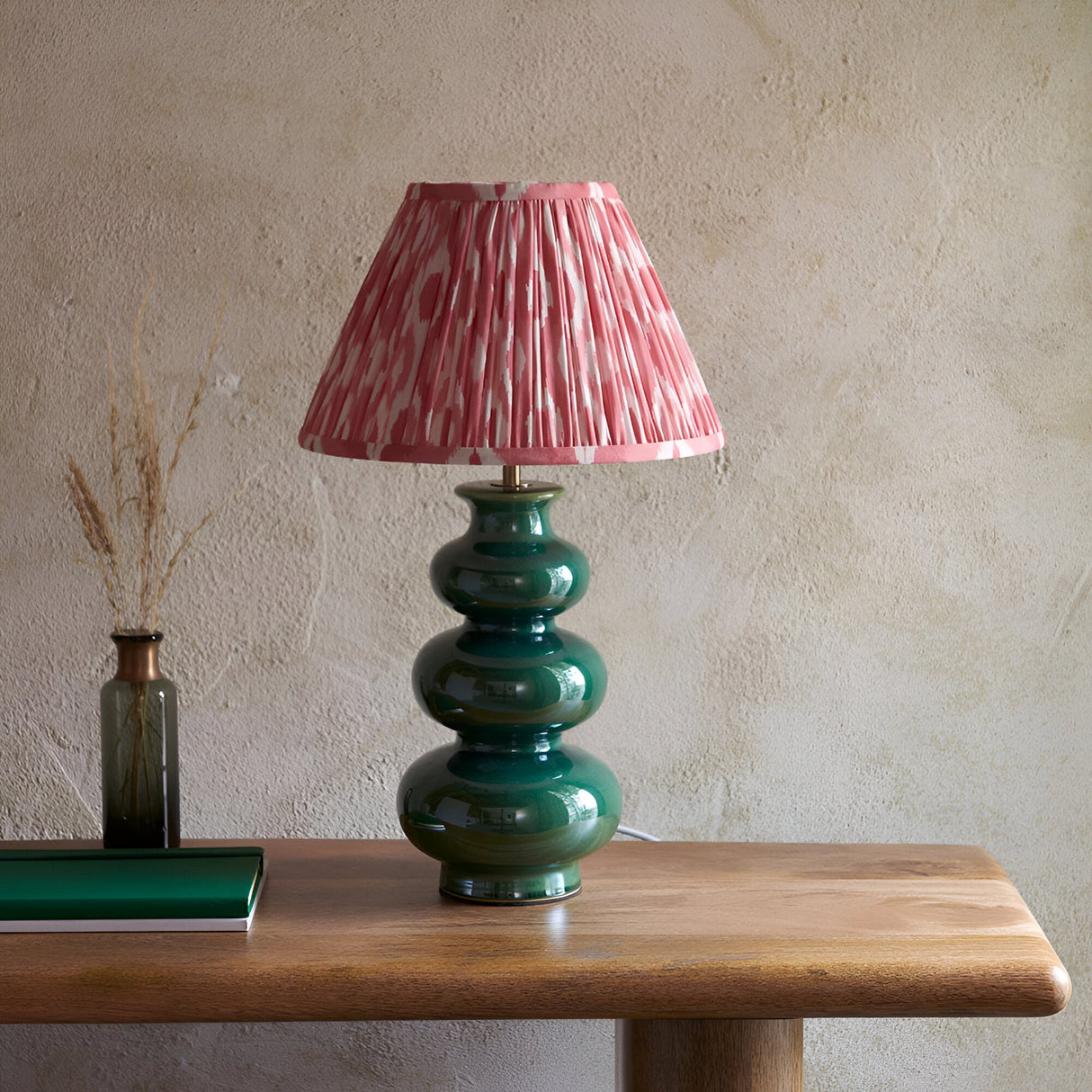 Carlstead Ikat Table Lamp with tiered glossy green ceramic base and pleated pink patterned shade, styled on a wooden table against a textured wall.