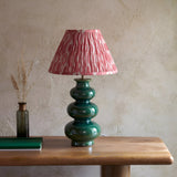 Carlstead Ikat Table Lamp with tiered glossy green ceramic base and pleated pink patterned shade, styled on a wooden table against a textured wall.