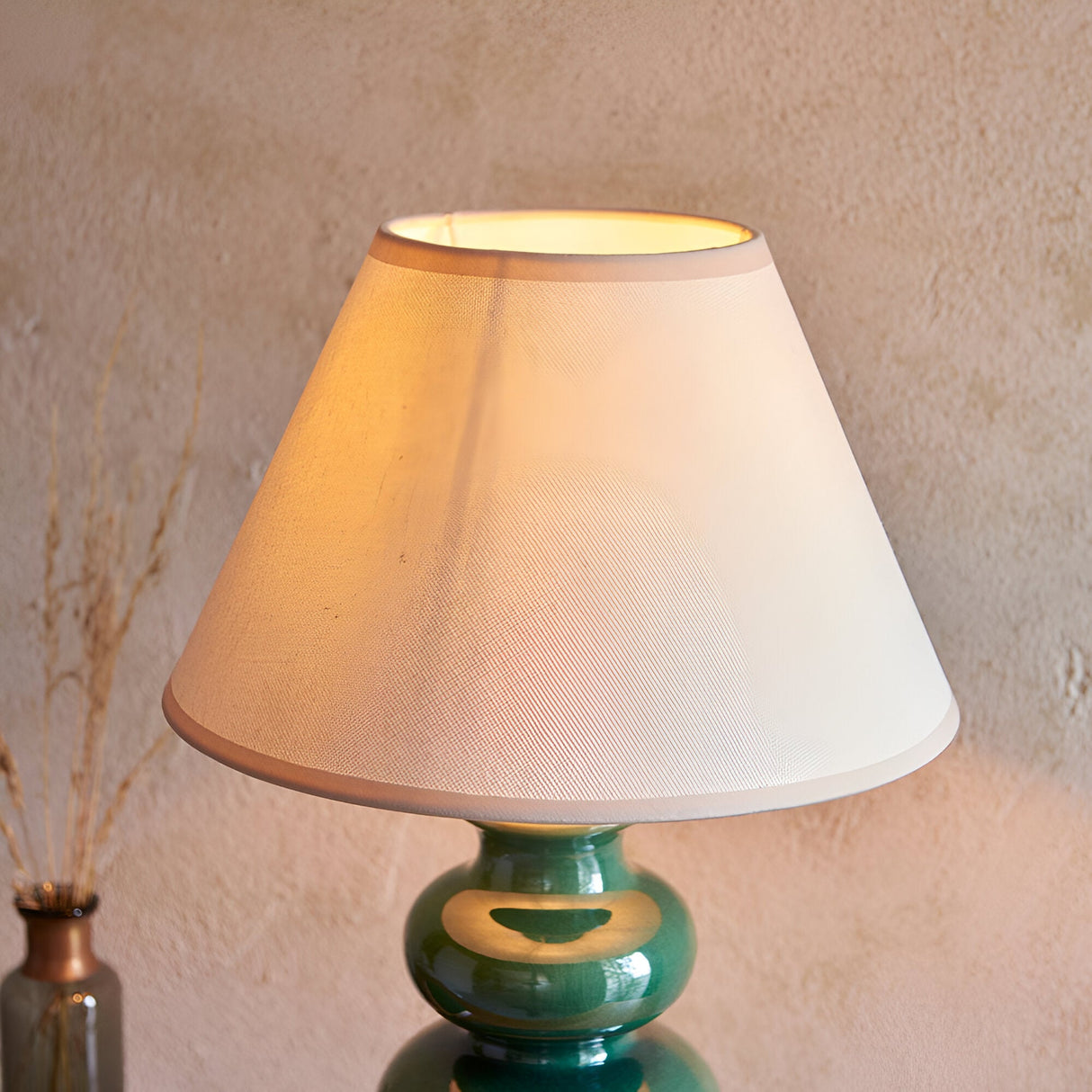 Green ceramic gourd-shaped table lamp with glossy reactive glaze and white cotton-mix shade, lit against a textured wall.