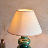 Green ceramic gourd-shaped table lamp with glossy reactive glaze and white cotton-mix shade, lit against a textured wall.