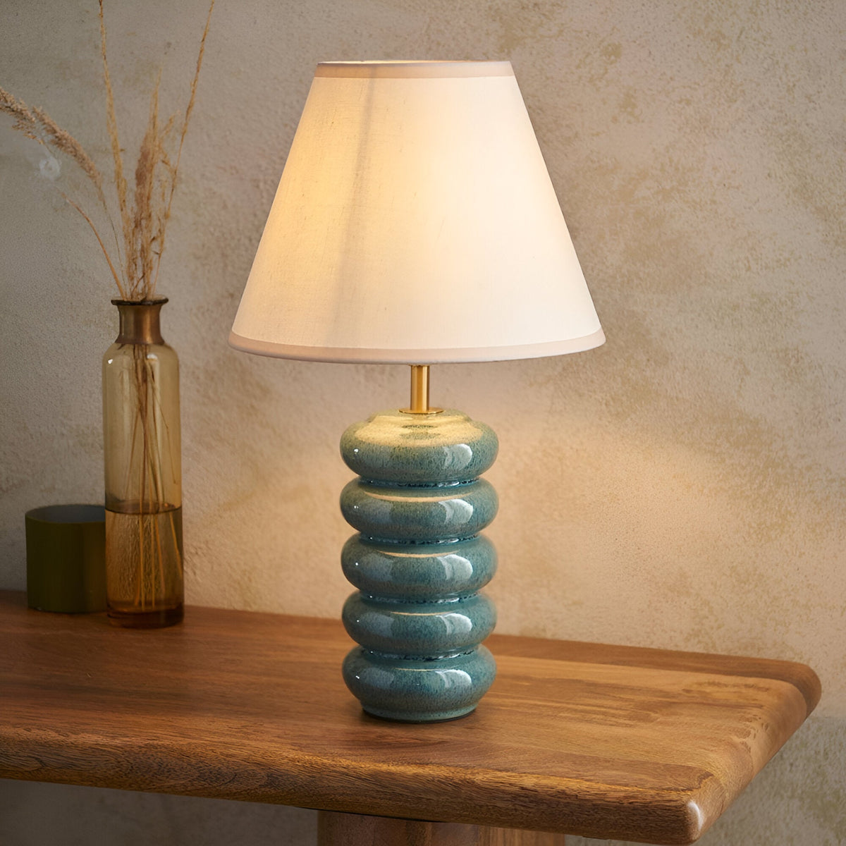 Endon Cliffstead table lamp with stacked ocean spray glazed ceramic base, antique brass stem and ivory tapered shade on a wooden console beside a glass vase.