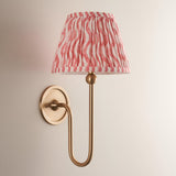 Endon Cliffleigh wall light with brushed aged brass curved arm and tapered hand-pleated coral pink Ripple faux linen shade, on a neutral background.