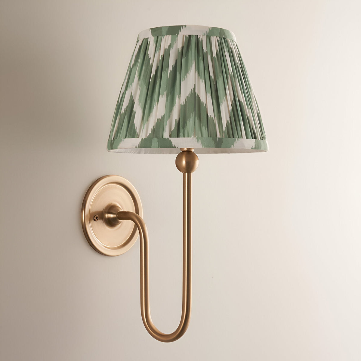 Endon Cliffleigh wall light with brushed aged brass curved arm and tapered hand-pleated green Zigzag faux silk shade, mounted on a neutral wall.