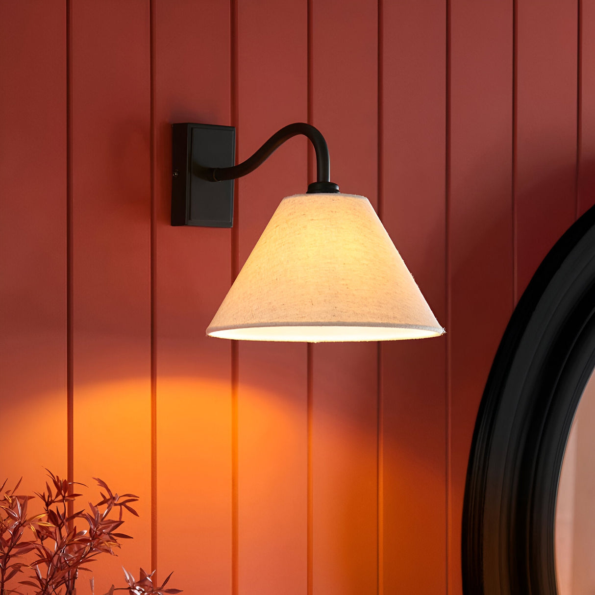 Endon Brockwood wall light with a curved matt black arm and tapered natural linen shade, mounted on a red panelled wall beside a round black-framed mirror.